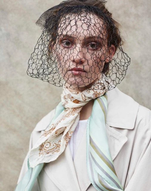 Close-up studio portrait of a woman wearing a mesh headpiece and patterned scarf, showcasing a fashion-forward aesthetic and neutral color palette.