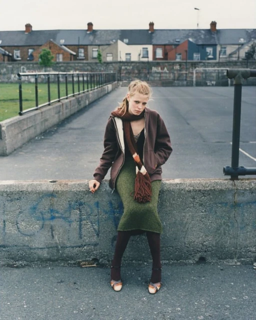 Young woman in brown jacket, olive skirt, and scarf sitting on concrete wall in urban setting, overcast day, looking serious.
