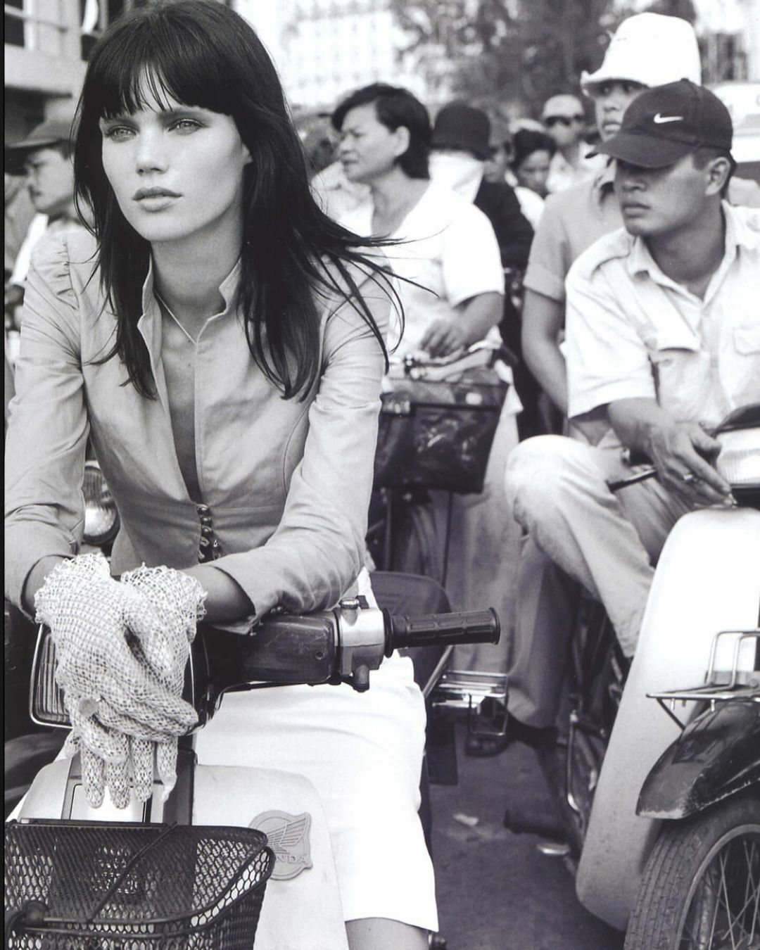 Monochrome shot of a woman riding a moped in an urban street scene. She wears gloves and a light-colored jacket, with other people and mopeds in the background.