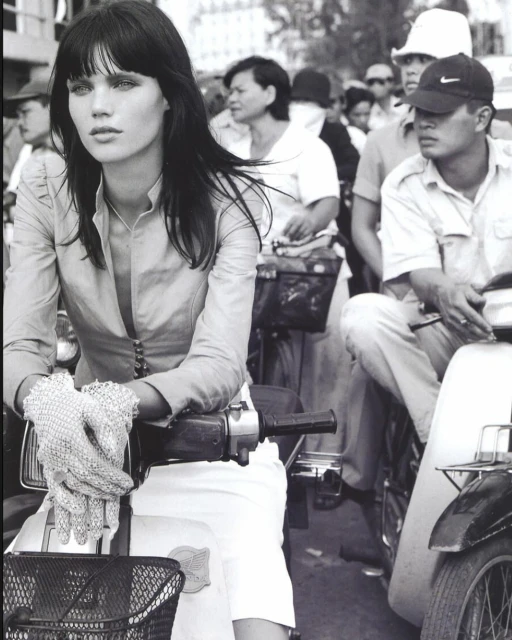 Monochrome shot of a woman riding a moped in an urban street scene. She wears gloves and a light-colored jacket, with other people and mopeds in the background.