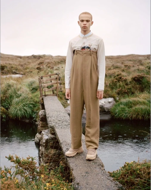 A person in oversized beige pants and an off-white top stands on a stone bridge, surrounded by a wild, grassy landscape under an overcast sky.
