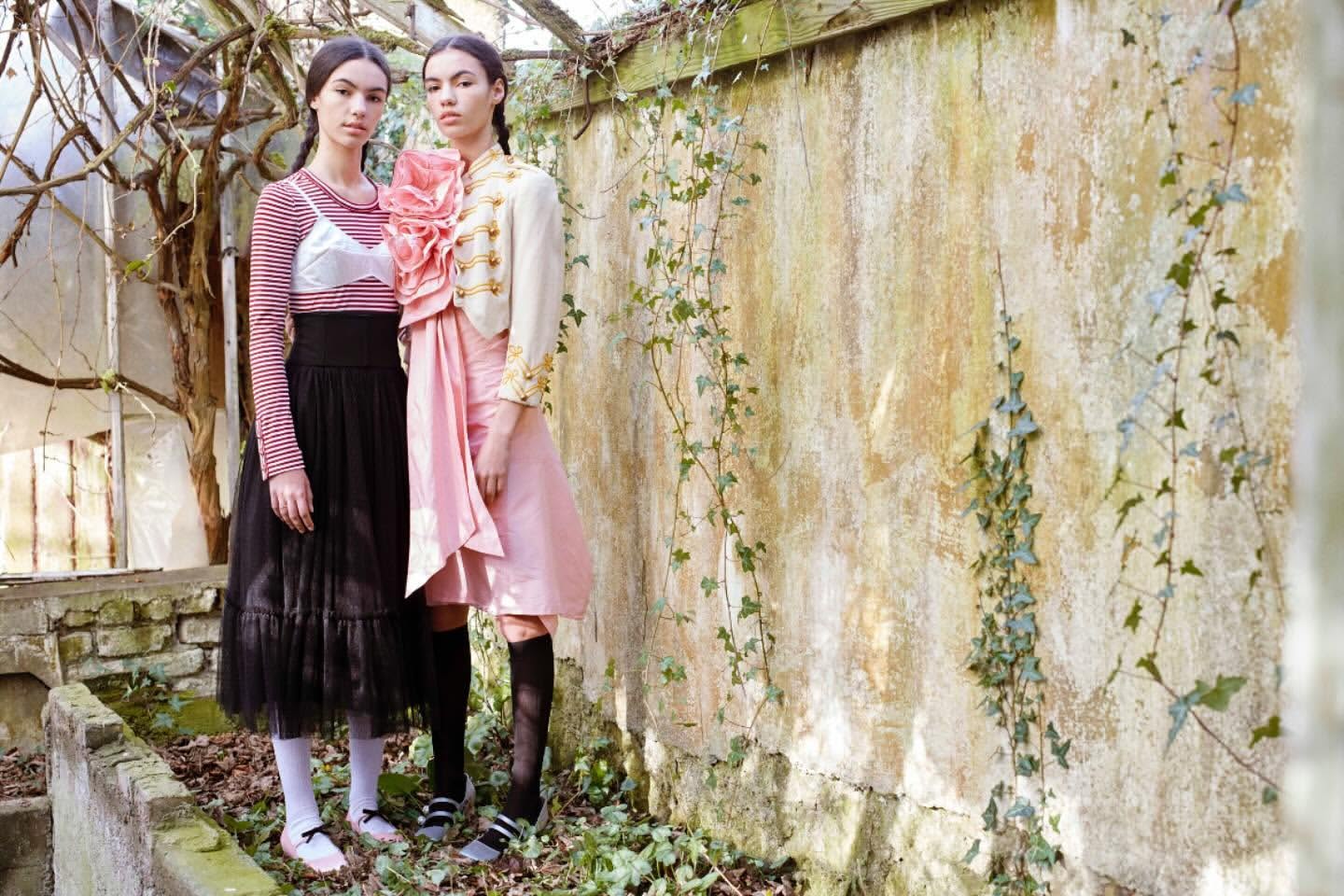 Two fashion models pose outdoors against a weathered wall covered in ivy. The women wear distinctive outfits in pink and black.