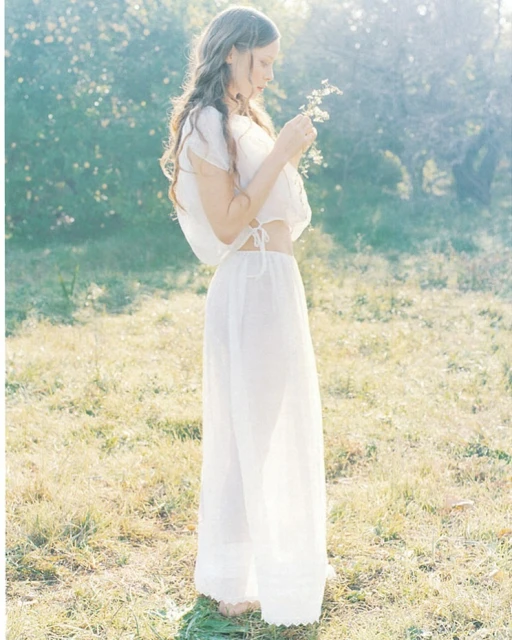 A young woman in white clothing standing in a field with wildflowers on a sunny day, creating a soft and ethereal atmosphere.