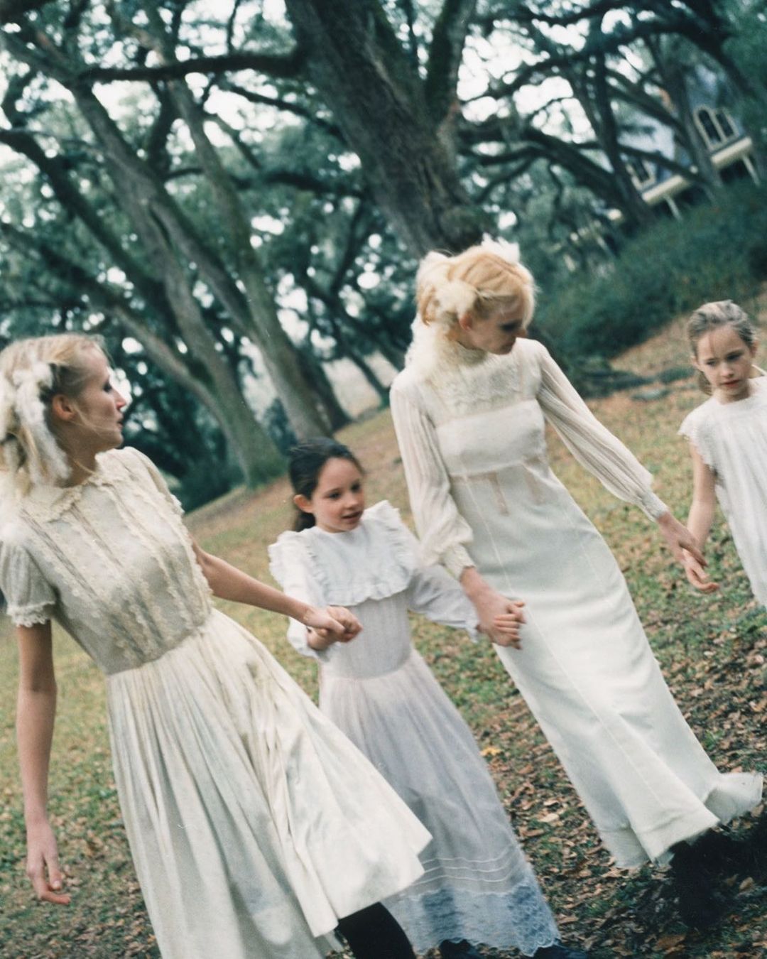 Four females in long white dresses holding hands in a park with large trees by photographer Perry Ogden.