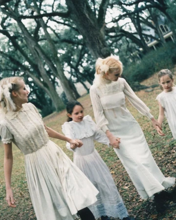 Four females in long white dresses holding hands in a park with large trees by photographer Perry Ogden.