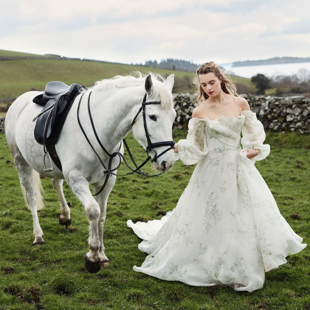 Woman in a white floral dress stands next to a white horse in a green field, with a stone wall and a distant body of water.