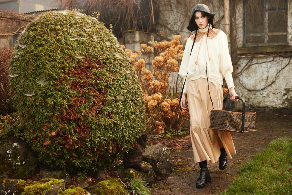 A woman in a long skirt and blouse stands on a path next to a mossy garden, carrying a brown tote bag in an outdoor fashion shot.