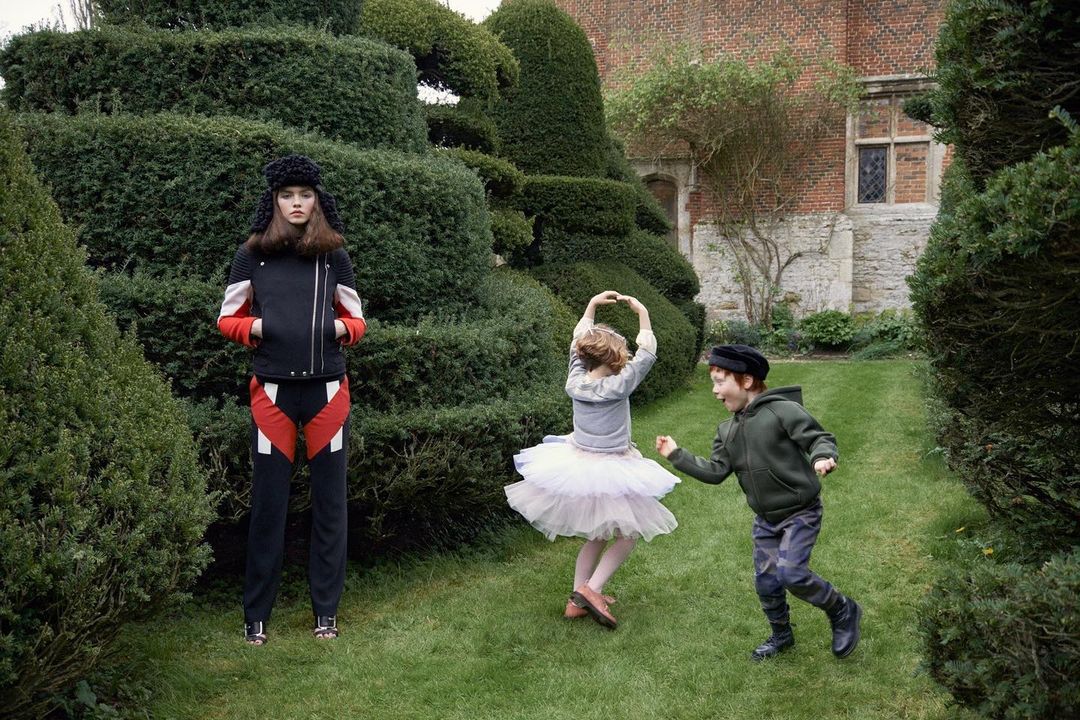 Fashion shot of a female model wearing a black jacket and pants, with two children playing in a topiary garden on a sunny day.