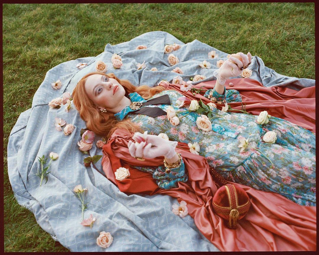 Red-haired woman in a floral dress lying on a light blue patterned blanket with pale pink roses scattered around on green grass outdoors.