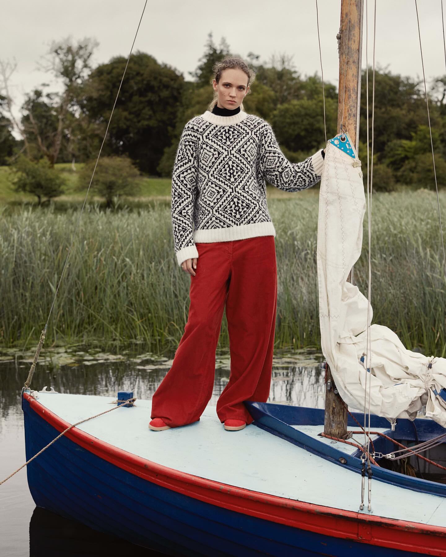 Fashion shot of a woman wearing a patterned sweater and wide-legged red pants, standing on a blue sailboat with a lake background.