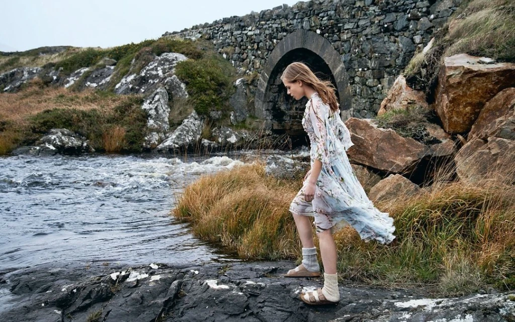 Young woman in floral dress walks on rocks near a river with a stone bridge in the background, blending fashion with a natural landscape.