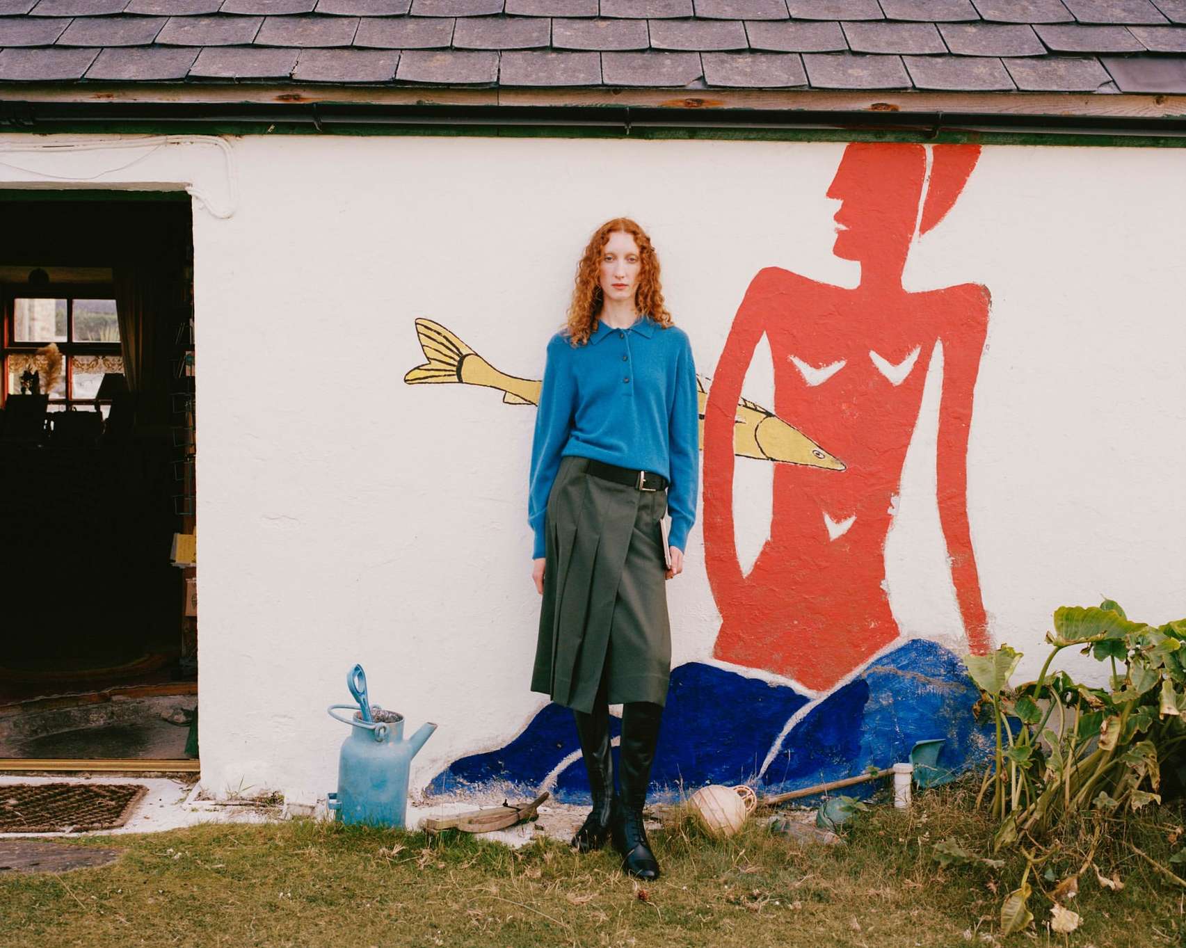 Red-haired adult woman in a teal sweater and dark green skirt, posing in front of an illustration on the white exterior wall of a cottage.