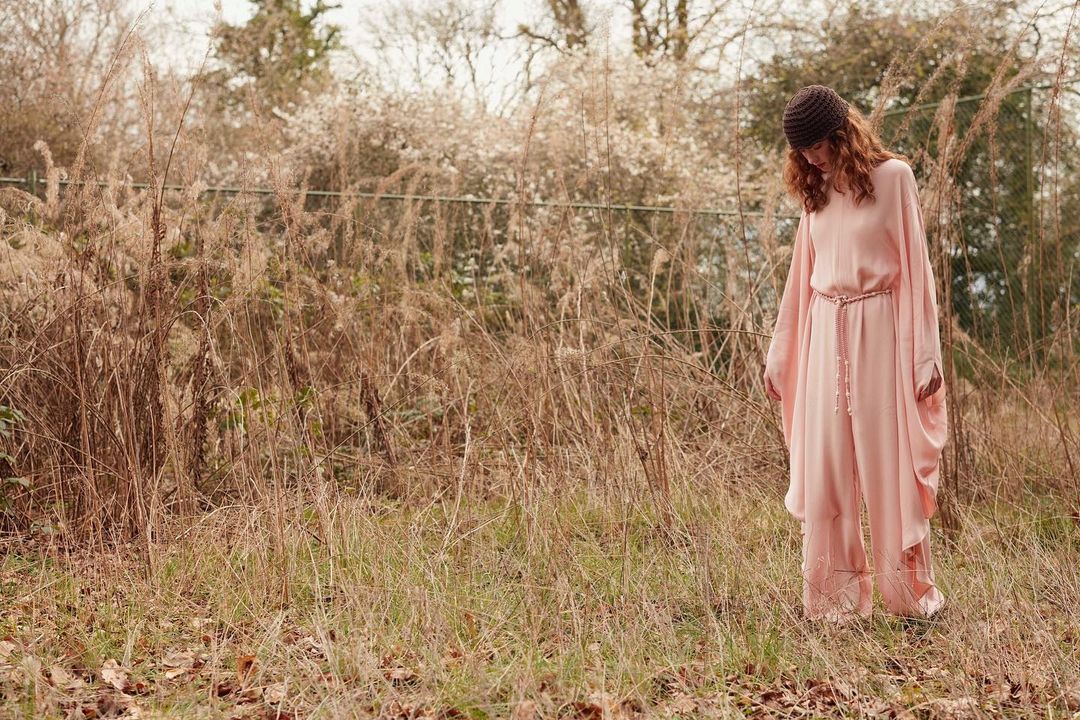 A woman wearing a pink jumpsuit and brown beanie stands in a field of tall, dry grass with trees and flowering bushes in the background.