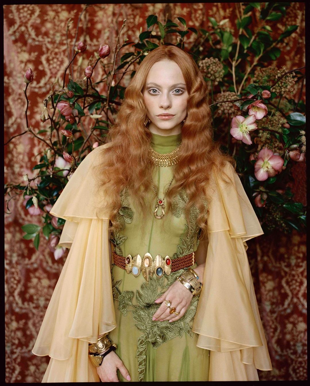 Red-haired woman in a flowing chartreuse dress and vintage gold jewelry poses in front of a floral tapestry and real flowers.