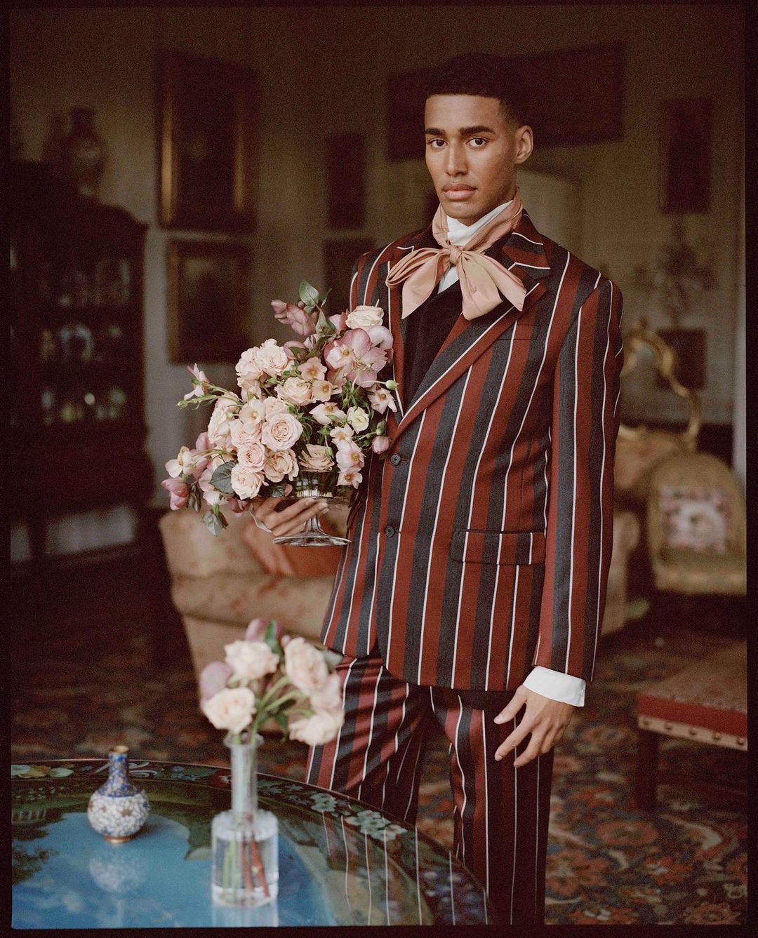 Portrait of a young adult male in a striped suit, holding a bouquet of flowers, in a grand interior setting with antique decor.