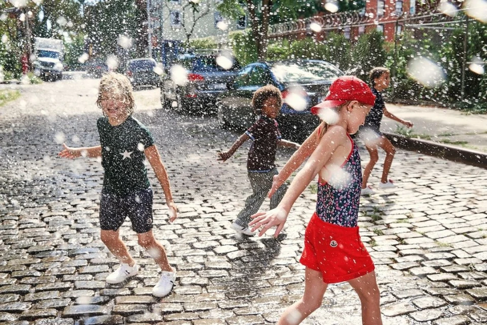 Group of children playing in water outdoors on a cobblestone street with cars and buildings in the background on a sunny day.