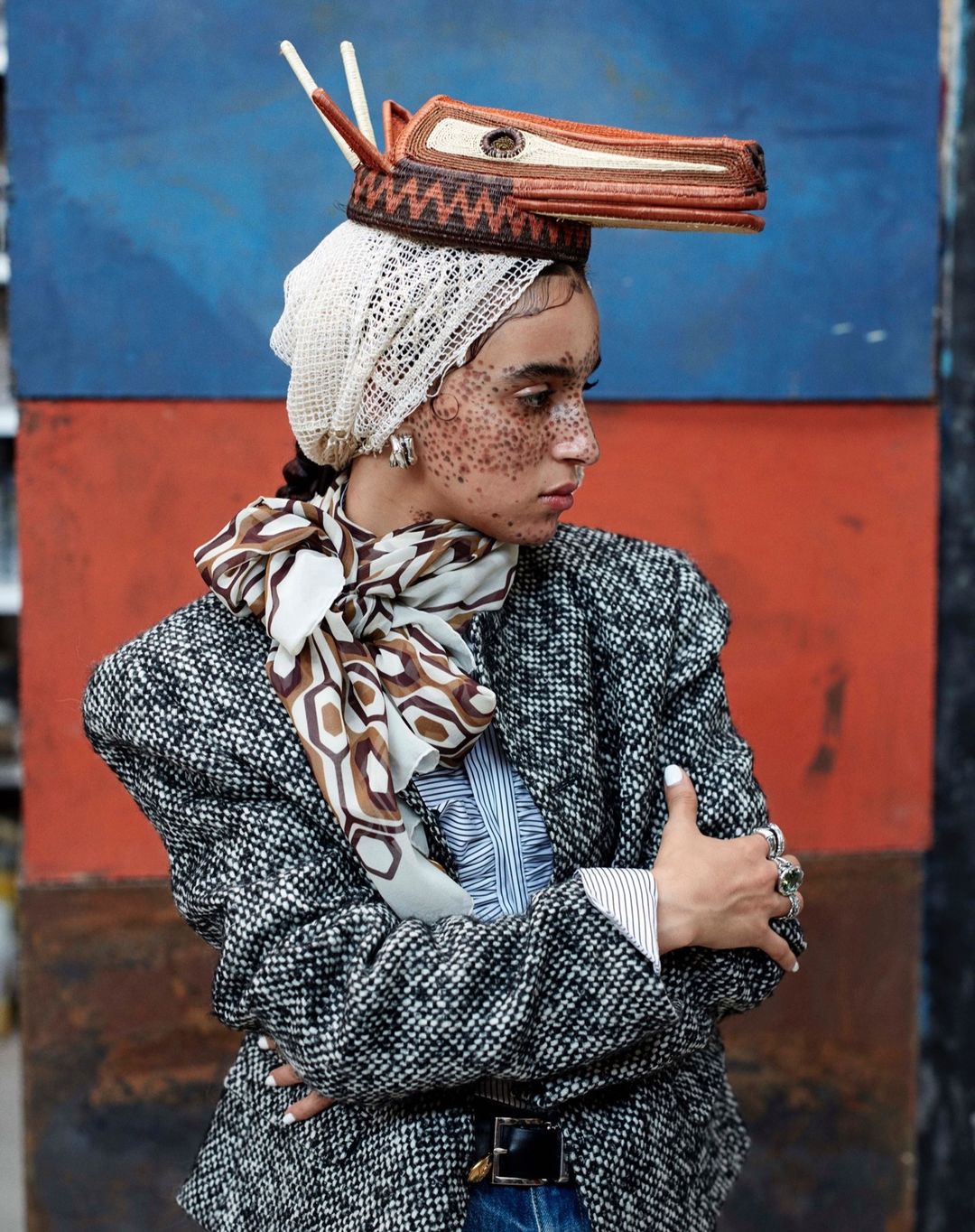 Fashionable woman wearing a tweed blazer, printed scarf, and unique headwear against a colorful textured wall backdrop in a studio setting.