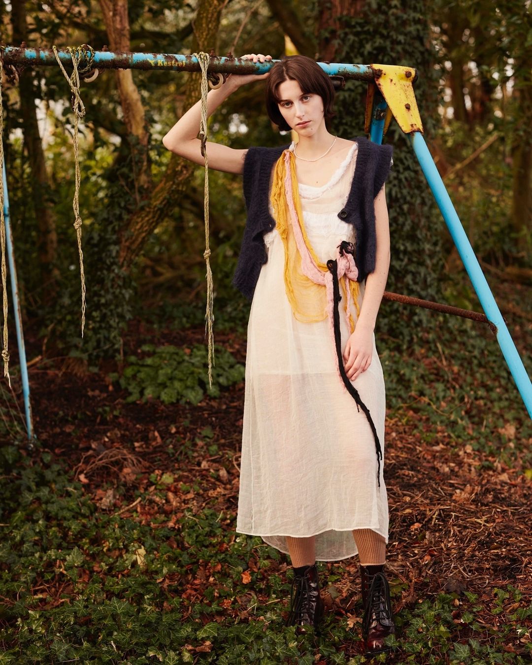 A woman in a white dress and dark vest poses next to a blue metal playground structure, set in a forest with leafy ground.
