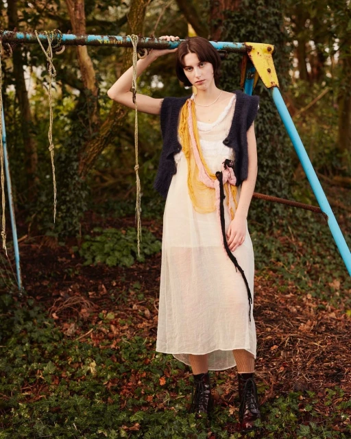 A woman in a white dress and dark vest poses next to a blue metal playground structure, set in a forest with leafy ground.