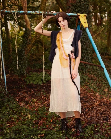 A woman in a white dress and dark vest poses next to a blue metal playground structure, set in a forest with leafy ground.