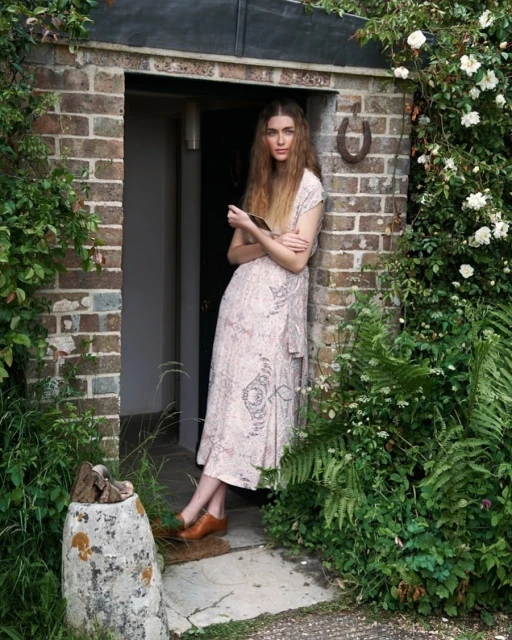 Woman in dress stands in doorway of brick building with horseshoe, surrounded by greenery and flowers in a rural setting.