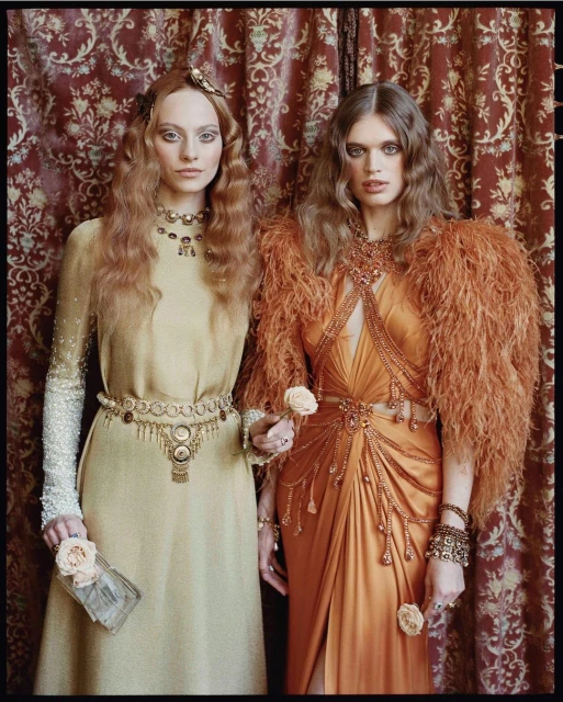 Two fashion models in retro-style gowns pose in front of a patterned curtain, holding small roses in a portrait fashion shot.