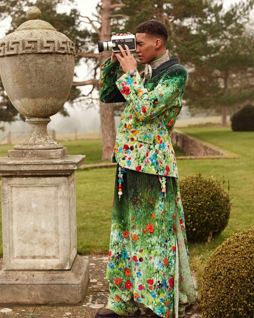 Man in floral suit films garden scene with vintage camera. Stone urn, green lawn, and trees in background. Fashion editorial photography.