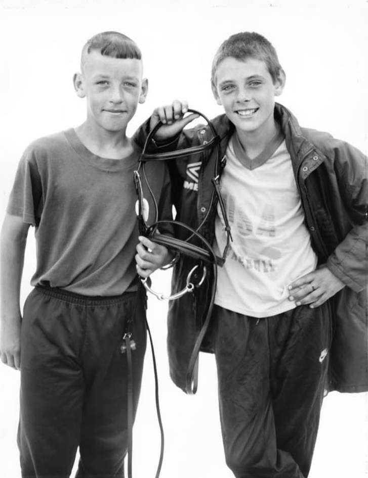 Two boys pose in a black and white photograph, one holding a horse bridle, wearing dark athletic wear and jackets on a bright day.