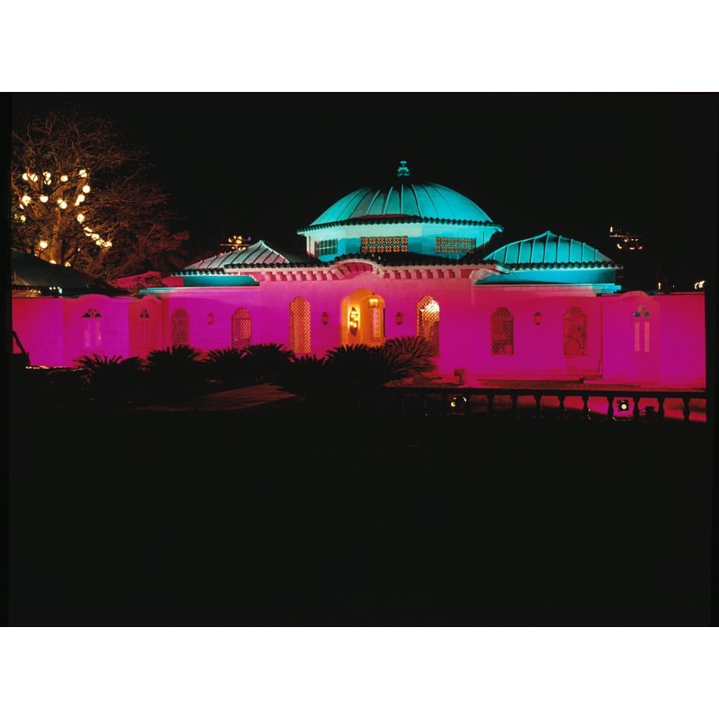 Nighttime view of an illuminated building featuring pink and turquoise lights, showcasing its architectural details and surrounding landscape.