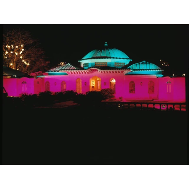 Nighttime view of an illuminated building featuring pink and turquoise lights, showcasing its architectural details and surrounding landscape.