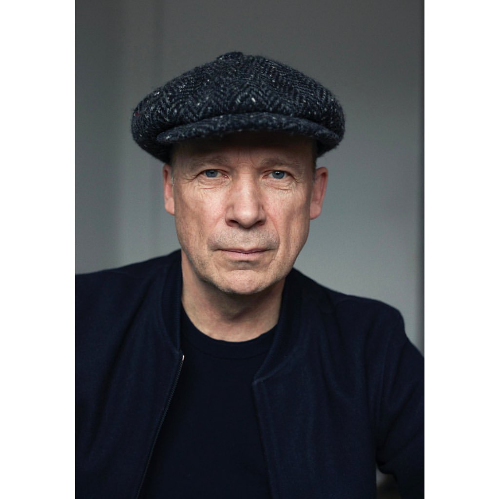 Portrait of an adult male wearing a navy blue jacket and a patterned newsboy cap, against a neutral gray background.