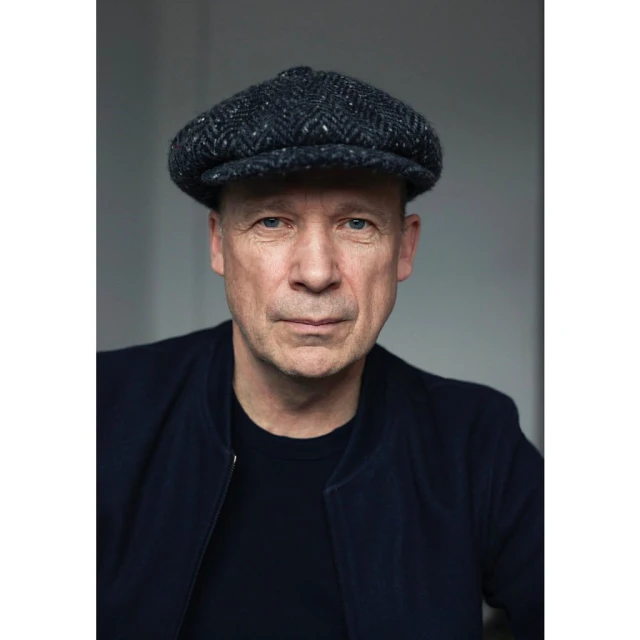 Portrait of an adult male wearing a navy blue jacket and a patterned newsboy cap, against a neutral gray background.