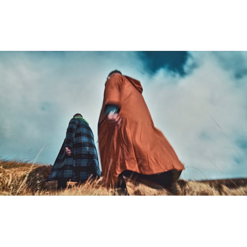 Two people walk on a grassy hill under a cloudy sky. One wears an orange hooded cloak, the other a blue and green plaid cloak.