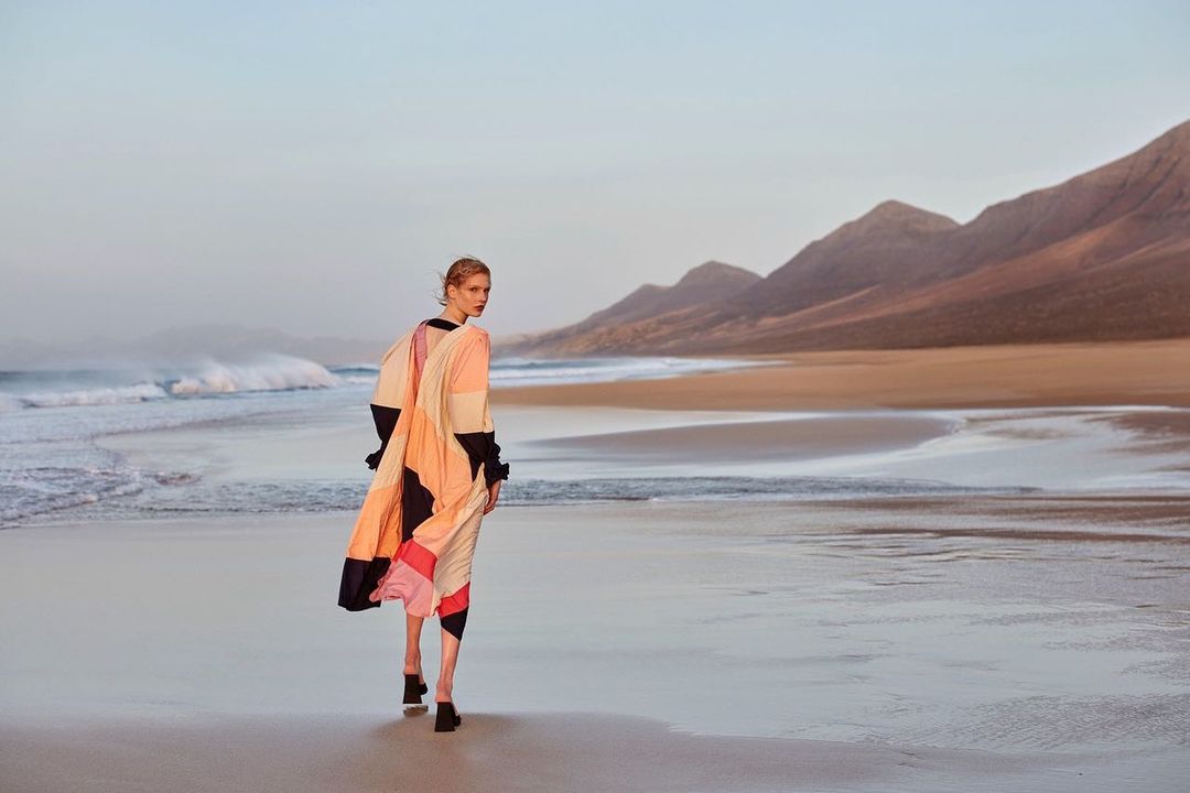 Fashion model with blond hair poses in patterned coat on sandy beach with distant mountains and ocean waves in outdoor fashion shot.