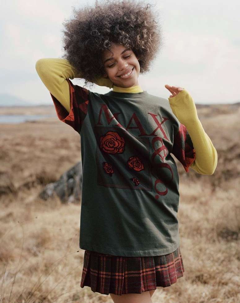 A woman with an afro smiles outdoors while wearing a yellow turtleneck, a large olive graphic tee, and a short, plaid skirt.