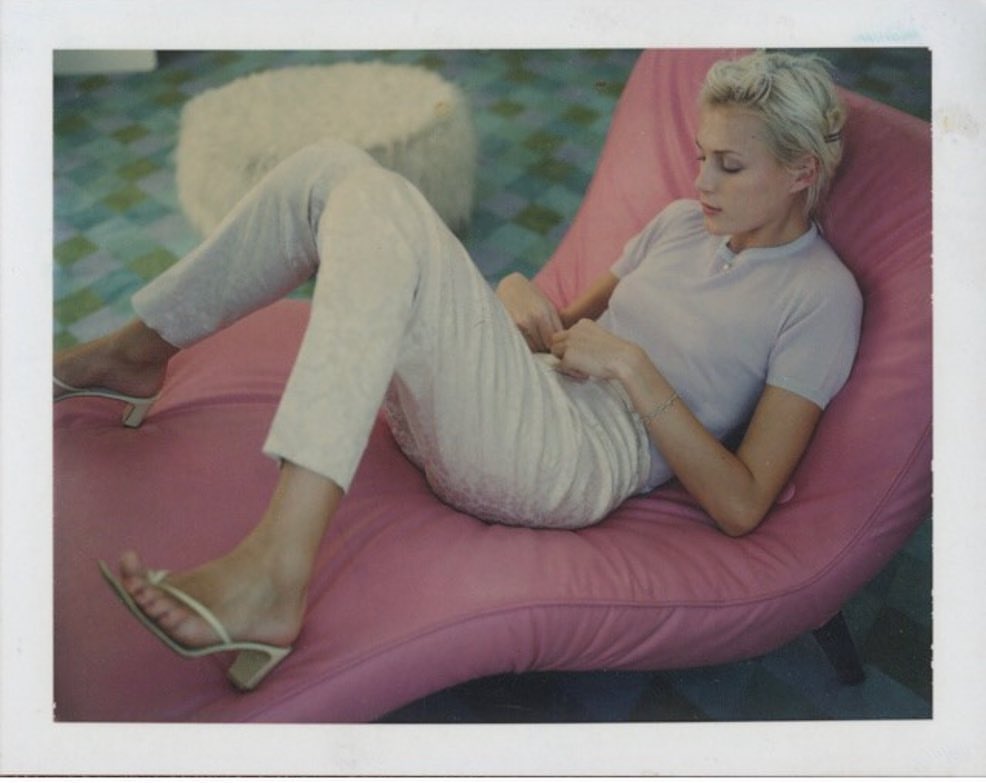 Woman lounging in a pink chair in a light-filled room with tile flooring. She wears pale pants and a light purple shirt, relaxing indoors.