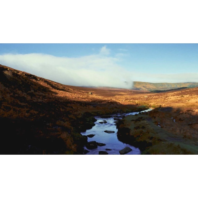 Scenic shot of a shallow stream flowing through a valley with sunlight on the hills and low-lying clouds in the sky on a sunny day.