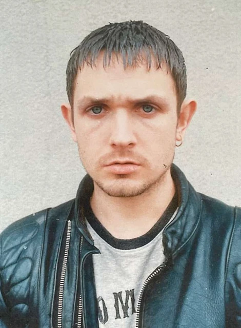 Close-up portrait of a young adult male with short hair, wearing a black leather jacket and grey t-shirt, against a grey wall.