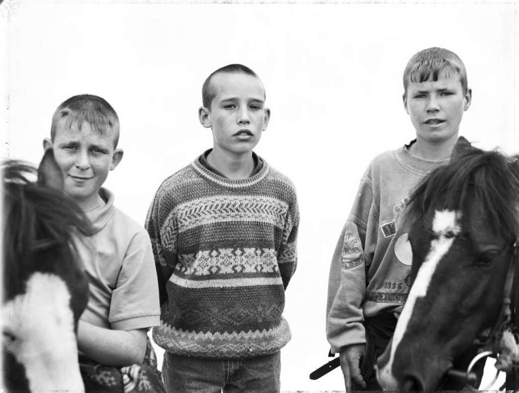 Black and white portrait of three young boys standing outside with two ponies, all facing forward, captured in a medium shot.
