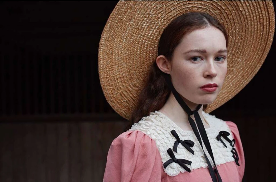 Portrait of a woman in a pink dress and straw sun hat with black ribbon ties, set against a dark wooden backdrop. Beauty and fashion.
