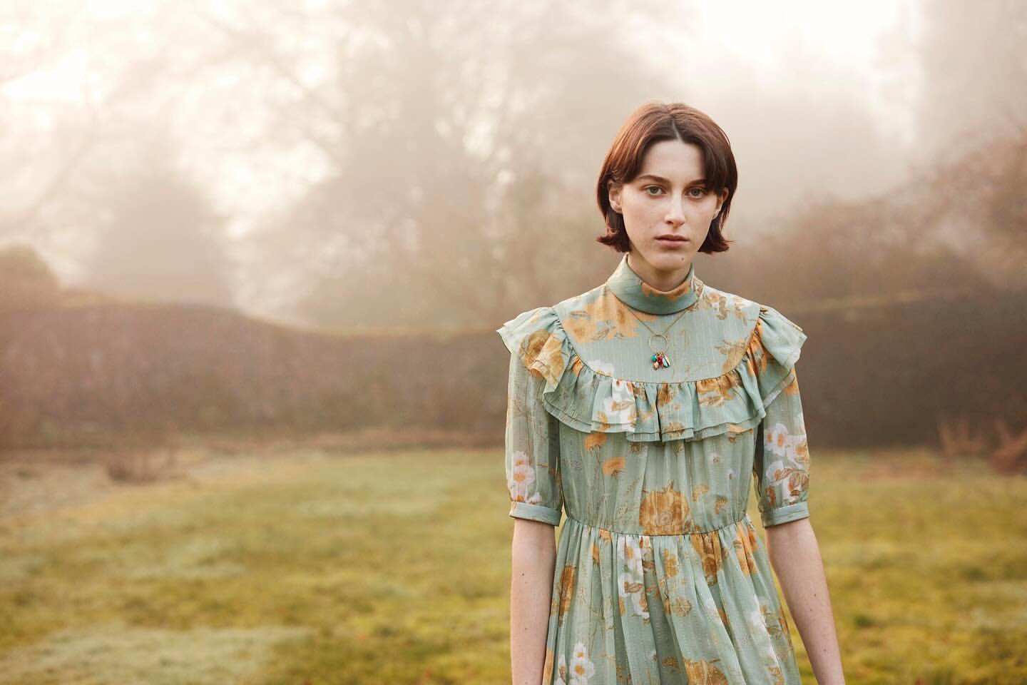 Portrait of a young adult woman in a floral dress standing in a grassy field with trees and a stone wall in the blurred, misty background.