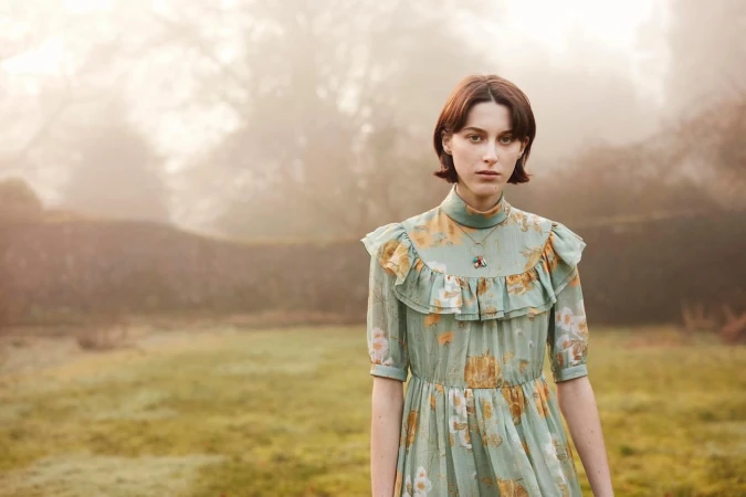 Portrait of a young adult woman in a floral dress standing in a grassy field with trees and a stone wall in the blurred, misty background.