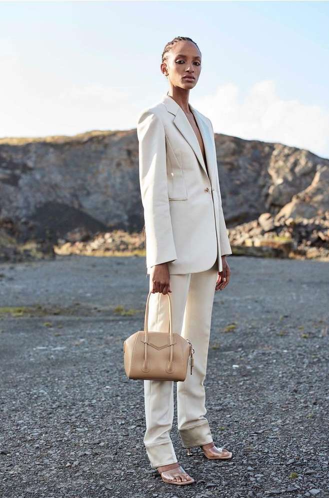 Woman in a cream suit with a purse standing against a rocky landscape. She wears clear heels and has her hair pulled back.