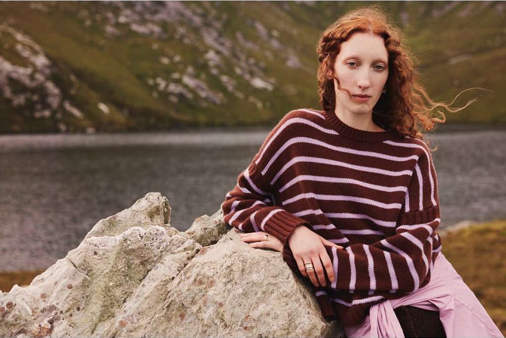 A red-haired woman in a striped sweater leans against a rock formation with a lake and mountains in the soft-focus background.