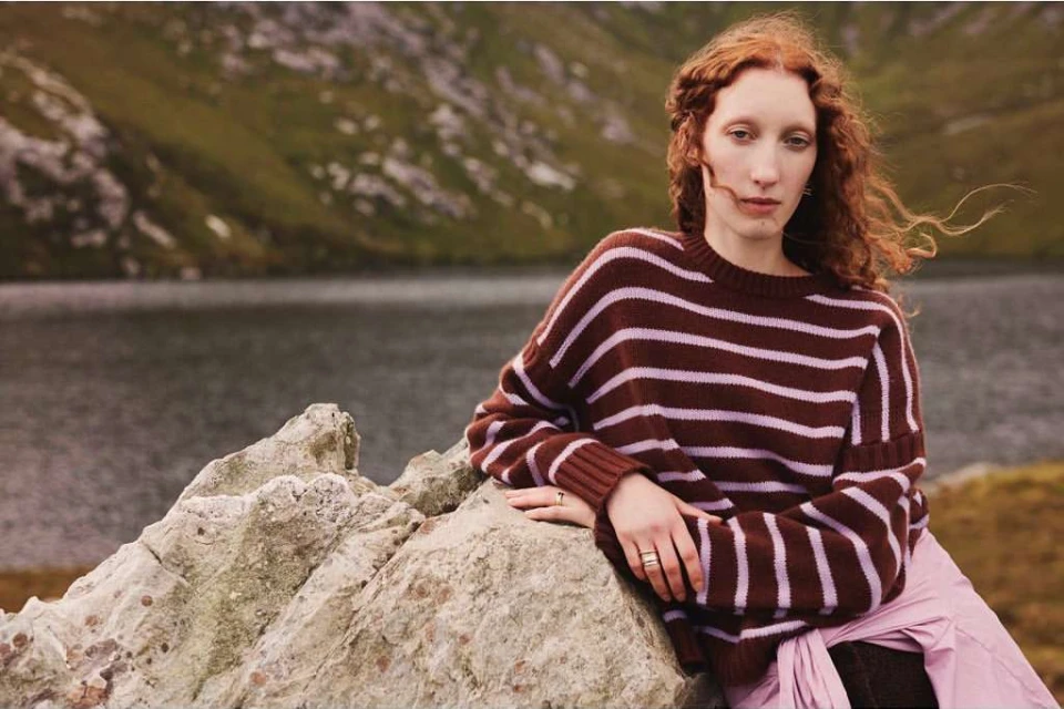 A red-haired woman in a striped sweater leans against a rock formation with a lake and mountains in the soft-focus background.