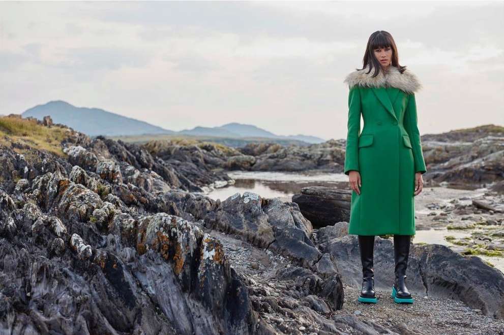A woman models a green coat with a fur collar on a rocky beach landscape under a cloudy sky in a fashion editorial shot.