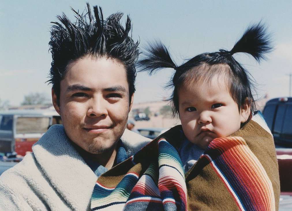 An adult male and an infant wrapped in a blanket are outdoors with buildings and parked cars visible in the background under a blue sky.