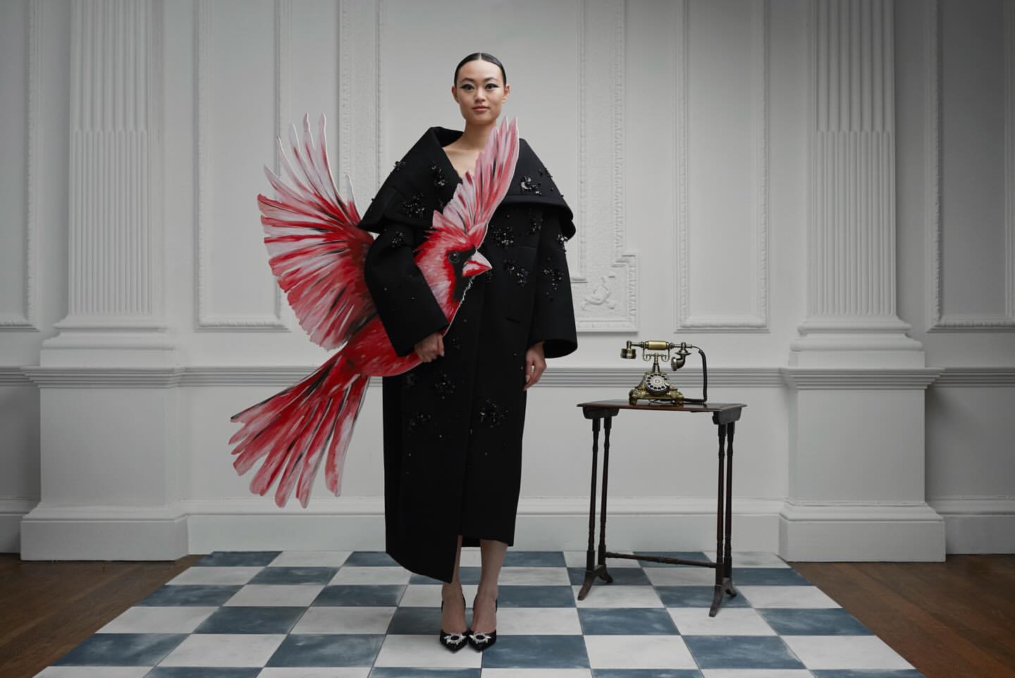 Asian woman in black dress holding red bird art, standing indoors on a blue-and-white checkered floor, next to an antique telephone.