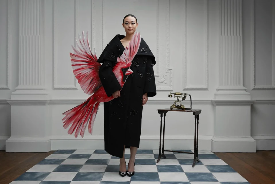 Asian woman in black dress holding red bird art, standing indoors on a blue-and-white checkered floor, next to an antique telephone.