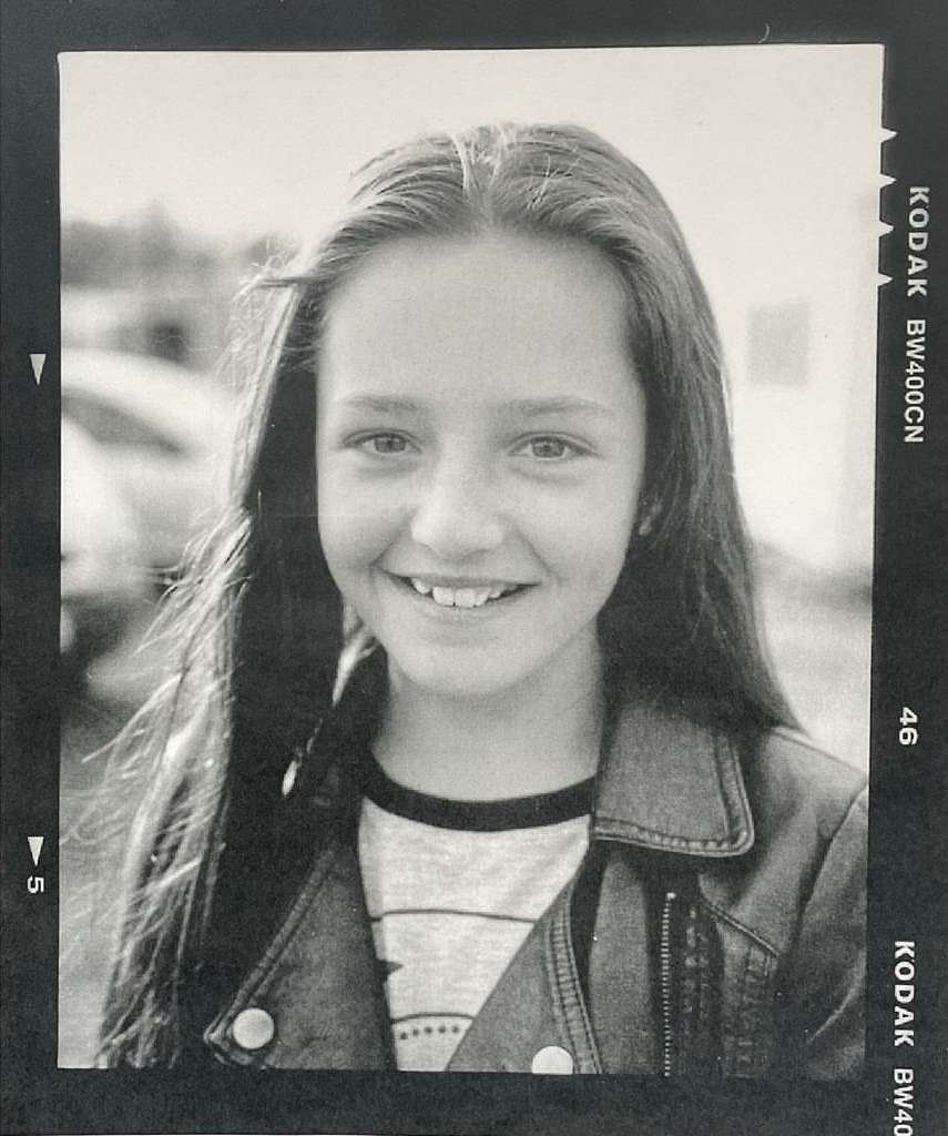 Black and white portrait of a smiling young girl with long dark hair wearing a leather jacket, framed by film border negatives.